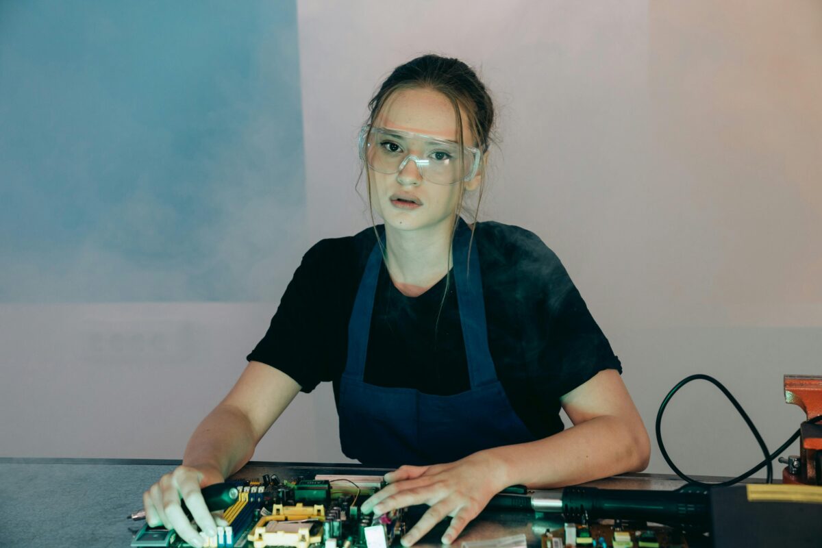 Woman in apron and goggles assembling electronic hardware indoors.